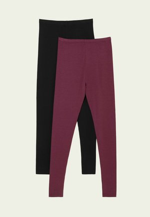 Mujer de pie con las manos detrás de la espalda, vestida con un cárdigan gris, leggings burdeos y zapatillas beige frente a un fondo claro y liso.