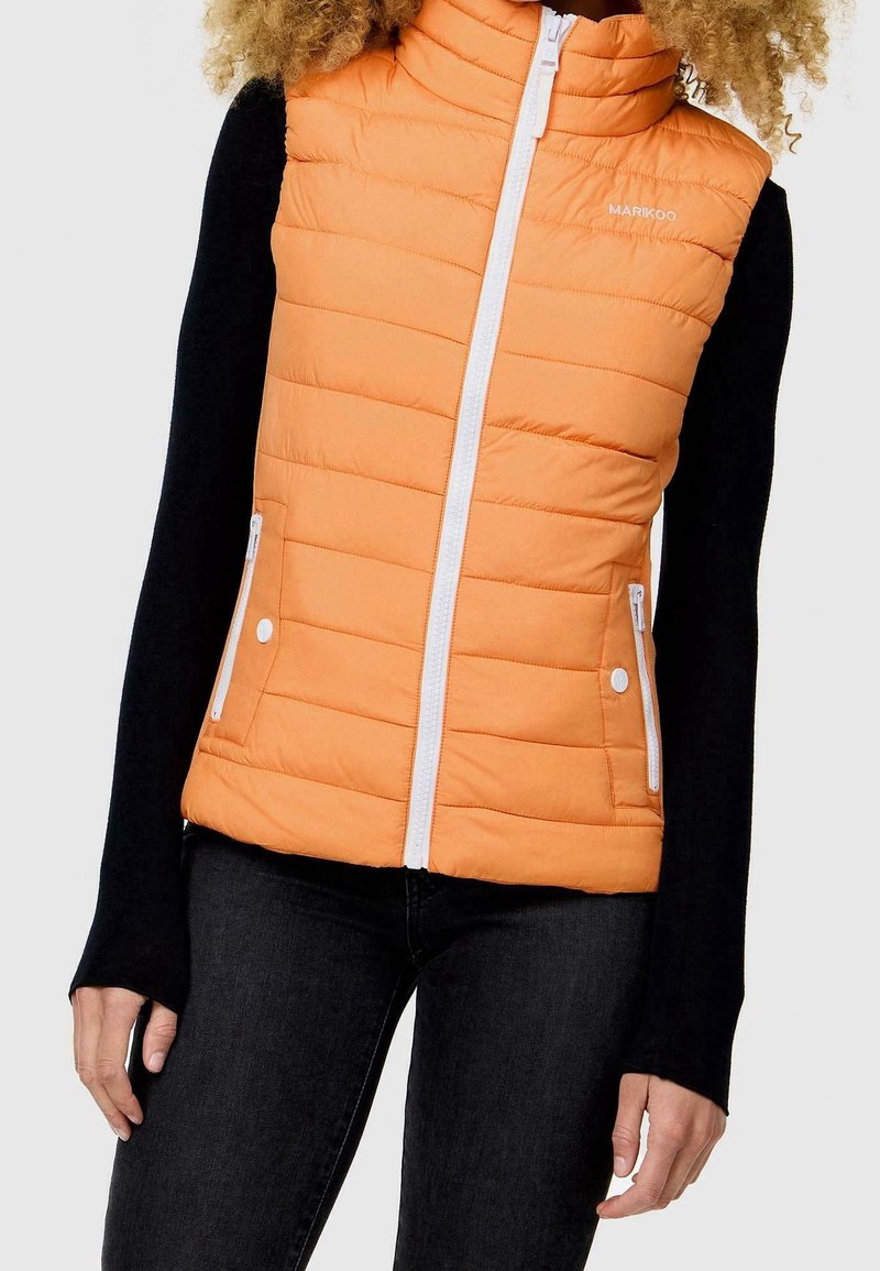 Femme portant un gilet matelassé orange sans manches avec une fermeture éclair blanche et des poches, par-dessus une chemise noire à manches longues et un pantalon noir.