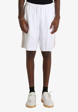 Persona che indossa pantaloncini bianchi con coulisse, calzini neri, sneakers bianchi con suola in gomma e un top nero, in piedi davanti a uno sfondo semplice.