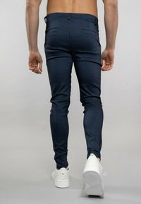 Modré přiléhavé kalhoty s texturovaným povrchem. Modelka má na nohou bílé tenisky a zezadu ukazuje štíhlou siluetu.