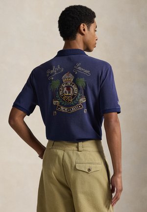Polo bleu foncé avec emblème brodé, présentant des palmiers et le texte "Polo Ralph Lauren". Associé à un pantalon kaki clair.