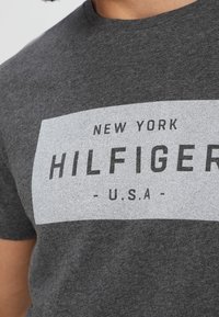 Grå kortærmet T-shirt med et kontrastfarvet hvidt rektangel, hvor "NEW YORK HILFIGER" og "U.S.A." er trykt med fede sorte bogstaver.