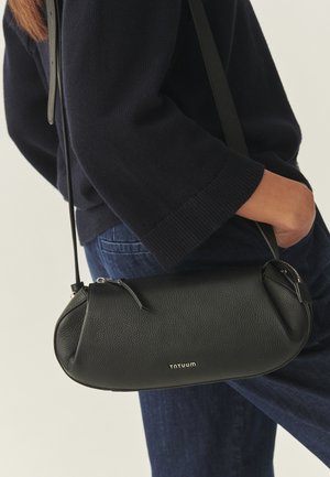 Sötét pulóvert és farmert viselő személy, aki egy Totuum logós fekete bőr oldaltáskát tart, keze a zsebében pihen.