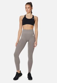 Leggings grigi alti in vita con una texture liscia abbinati a un reggiseno sportivo nero con logo. Modella in piedi con sneaker nere.