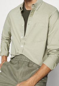 Vaaleanvihreä napitettu paita, joka on valmistettu puuvillasta, rintataskulla ja käännetyillä hihoilla, yhdessä oliivinvihreiden jousto- vyötäröhousujen kanssa.