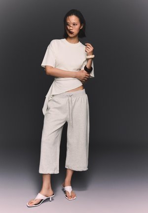 Donna in piedi contro uno sfondo scuro che indossa una maglietta bianca a maniche corte, pantaloni larghi grigio chiaro e sandali bianchi con tacco, con due braccialetti.