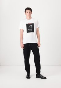 Hvit t-skjorte med korte ermer og svart hai-grafikk samt "HUGO"-tekst, sammen med svarte joggebukser og svarte dressko.