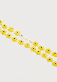 Des perles en forme de fleurs jaunes avec des centres bleus et de petites perles d'espacement blanches forment un bracelet avec un fermoir en argent sur un fond blanc.