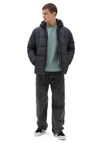 Padded czarna kurtka z kapturem, zamek błyskawiczny i pikowany wzór, w połączeniu z jasnoniebieską bluzą i ciemnoszarymi dżinsami. Sneakersy mają czarne i białe akcenty.