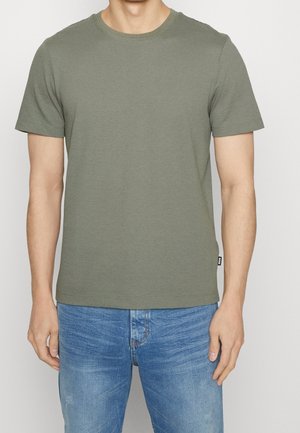 Homme portant un t-shirt vert olive texturé à manches courtes et un jean bleu clair, debout devant un fond uni.