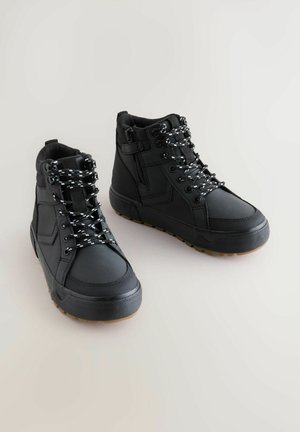 Svart höga sneakers har texturerade sidor, svarta och vita snören, en sidodragkedja och en gummisula med ett brunt mönster.