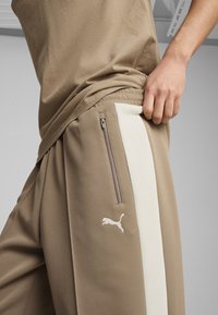 Brune atletiske bukser med en hvid sidestribe, lynlåslomme og et lille hvidt logo på låret. Stoffet ser struktureret og letvægtigt ud.