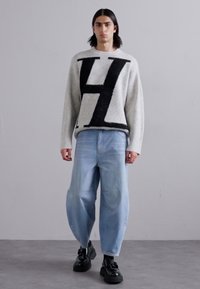 Γκρίζα μάλλινη ζακέτα με έντονο μαύρο σχέδιο "Η", συνδυασμένη με ανοιχτό μπλε φαρδιά τζιν. Το μοντέλο φοράει μαύρα παπούτσια με υφή.