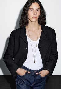 Osoba s kudrnatými tmavými vlasy, oblékající černou texturovanou bundu, bílý top bez rukávů, vrstvené náhrdelníky a modré džíny, s rukama v kapsách.