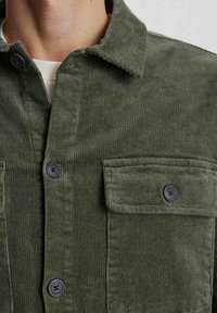 Cămașă din corduroy verde măsliniu cu guler ascuțit, butoni negri și un buzunar pe piept. Material texturat și detalii cu dungi verticale.