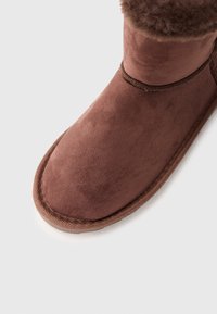 Brune ankel støvler i semsket lær med myk pelskant øverst. Rund tå, polstret såle og synlige sømmer langs kanten.