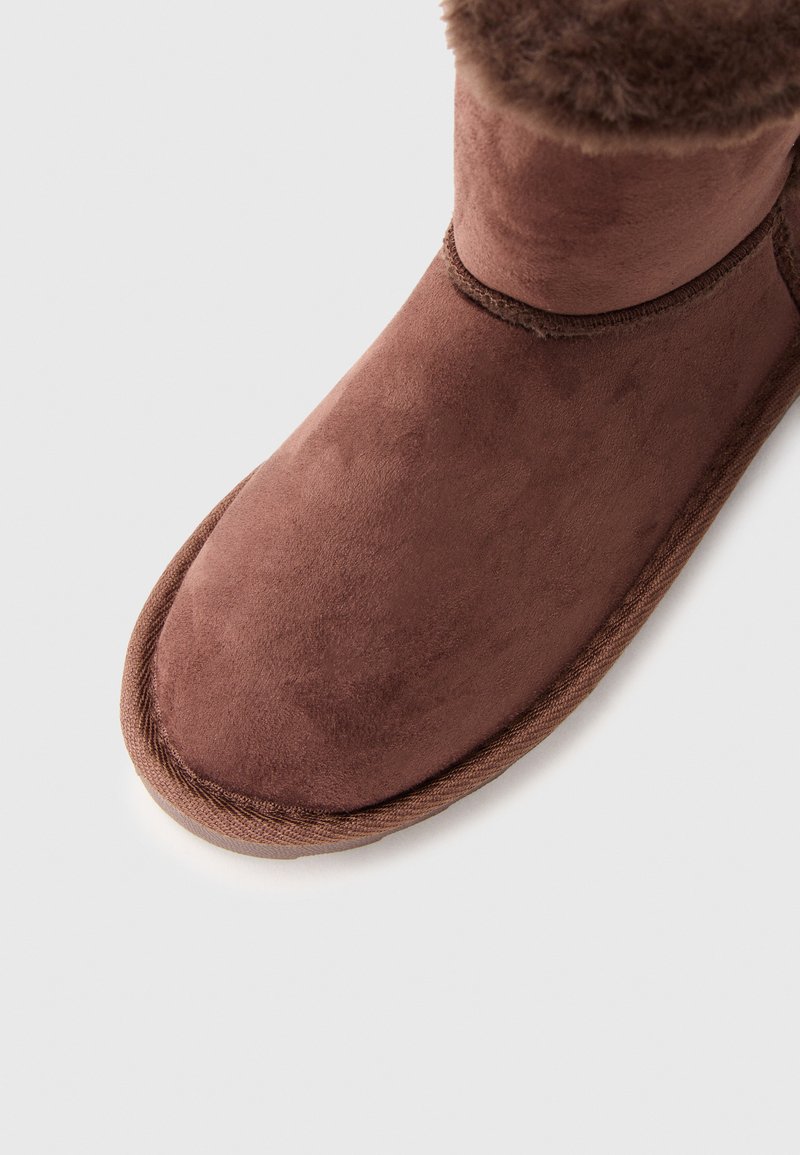 Bruine suède enkelboot met een zachte bontafwerking aan de bovenkant. Rond neus, gevoerde zool en zichtbare stiksels langs de rand.