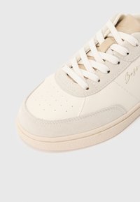 Balta sporta kurpe ar gludu ādas augšdaļu, zamšādas akcentiem, perforētu purngala daļu un bēša krāsas šņorēm. Sānos ir smalks zīmola uzraksts.