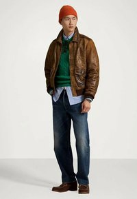 Jeune homme portant une veste en cuir marron, un pull vert, une chemise bleue, un jean noir, des bottes marron et un bonnet orange, se tenant avec les mains dans les poches.