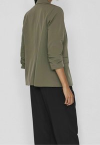 Blazer vert olive avec un col cranté, manches trois-quarts et coupe décontractée, associé à un pantalon noir. Texture de tissu lisse.