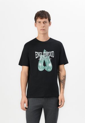Young man wearing black t-shirt with green boxing gloves graphic and "Englewood Boxing Gym" text, paired with gray pants, standing against white.