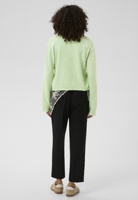 Pull-over vert clair, coupe courte, associé à un pantalon noir avec des détails en dentelle à l'arrière. Chaussures beige complètent la tenue.