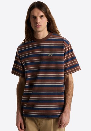 Ung mand med langt hår iført en brun og navy stribet Vans t-shirt og beige bukser, stående mod en ensfarvet baggrund.