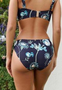 Femeie îmbrăcată într-un bikini bleu închis cu un model floral deschis, fotografiată din spate cu un fundal de grădină.