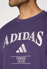 Lila pamut póló, rajta fehér "ADIDAS" logóval, alatta a "TENNIS CLUB" felirattal és a jellegzetes háromcsíkos emblémával. Puha tapintású, kerek nyakú.