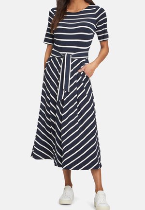 Femme portant une robe midi bleu marine avec des rayures blanches horizontales sur le haut, des rayures diagonales sur la jupe, une ceinture à la taille, et des baskets blanches.