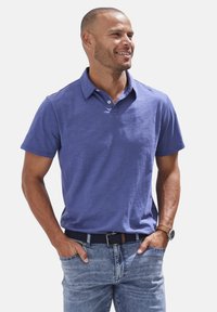 Lila Poloshirt mit kurzen Ärmeln und knöpfbarem Kragen, aus strukturiertem Stoff, kombiniert mit hellblauen Jeans und einem dunklen Gürtel.