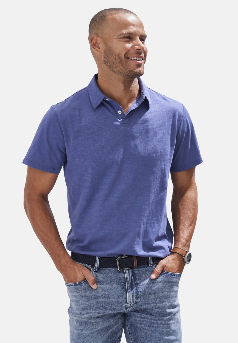 Lila Poloshirt mit kurzen Ärmeln und knöpfbarem Kragen, aus strukturiertem Stoff, kombiniert mit hellblauen Jeans und einem dunklen Gürtel.