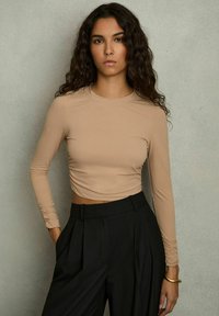 Hu nude met lange mouwen en een cropped model, voorzien van geplooide details bij de polsen, gecombineerd met hoge zwarte broeken en een gouden armband.