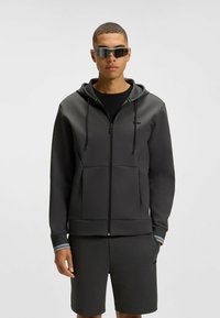 Felpa zip grigia con cappuccio, con accenti verdi, abbinata a pantaloni corti grigi. Il modello indossa occhiali da sole riflettenti, su uno sfondo chiaro.