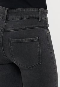 Tesne prilegajoči črni jeans s prikazanim zadnjim žepom, zankami za pas in delom pasu na svetli podlagi.