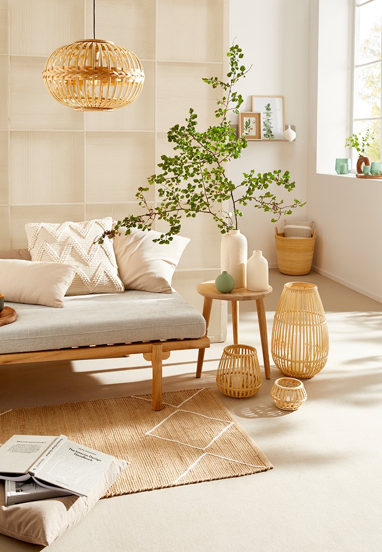 Holzsofa mit grauen Kissen, geflochtene Beleuchtung, runder Beistelltisch, dekorative Vasen und ein Teppich aus Naturfasern. Grünes Pflanzenakzent.