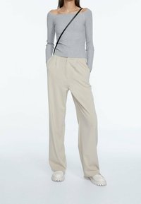 Kobieta w szarej bluzce na jedno ramię z prążkowego materiału, beżowych szerokich spodniach, białych grubych butach i czarnym pasku torby na ramię.