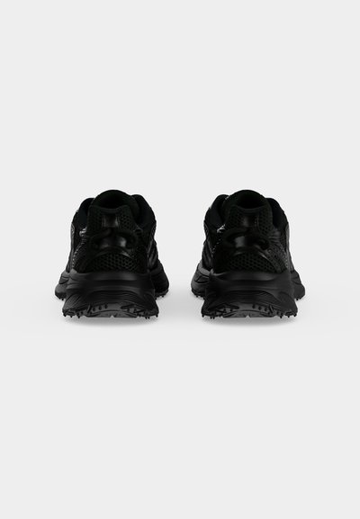 Paire de chaussures de sport noires vues de dos sur un fond clair uni, montrant des semelles crantées et des détails en mesh.