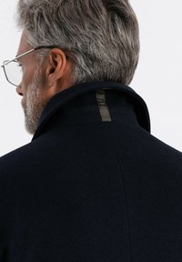 Cappotto di lana blu navy con colletto alto, caratterizzato da un sottile dettaglio a loop sul retro e una texture liscia, visto da un'angolazione posteriore.
