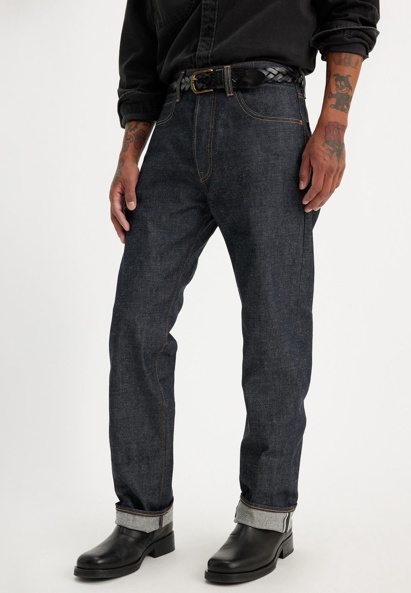 Homme portant un jean foncé retroussé avec des coutures marron, une ceinture tressée noire, une chemise noire, des bottes noires et des avant-bras tatoués.