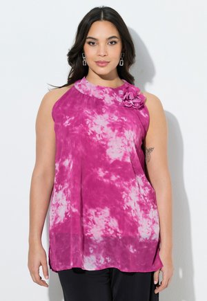 Frau mit langen dunklen Haaren trägt ärmelloses, pink-weißes Batik-Oberteil mit Blumendetail an der Schulter, schwarze Hose, steht vor weißer Wand.