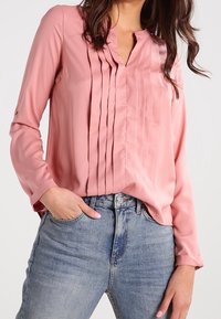 Blouse en soie rose avec plis verticaux, design sans col, manches longues et détails à boutons. Associée à un jean bleu délavé.