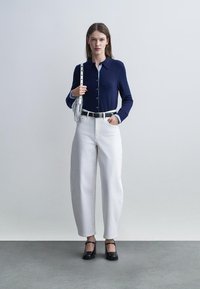 Frau steht und trägt einen marineblauen, geknöpften Pullover, weiße, hoch taillierte Weitbein-Hosen, schwarze High Heels und eine weiße Schultertasche mit Fransen.