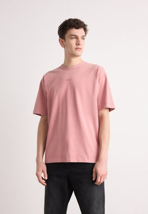 Light pink cotton t-shirt with short sleeves, round neck, and subtle logo in the center. Worn with black denim jeans.