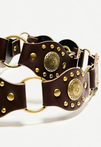 Brauner Ledergürtel mit runden goldenen Medaillons und Nieten, mit in das Design integrierten Metallringen. Strukturierte Oberfläche sichtbar.