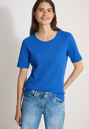 Femme souriante aux longs cheveux foncés portant un t-shirt bleu vif à manches courtes et un jean bleu clair, debout devant un fond uni.