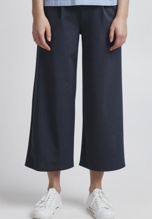 Person trägt marineblaue weite, gekürzte Hosen und weiße Sneakers, steht vor neutralem Hintergrund.