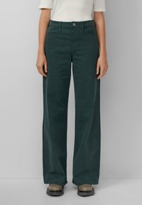 Dunkelgrüne, weit geschnittene Hose aus glattem Material mit einem Knopfverschluss vorne und Gürtelschlaufen. Kombiniert mit braunen Plateau-Schuhe.