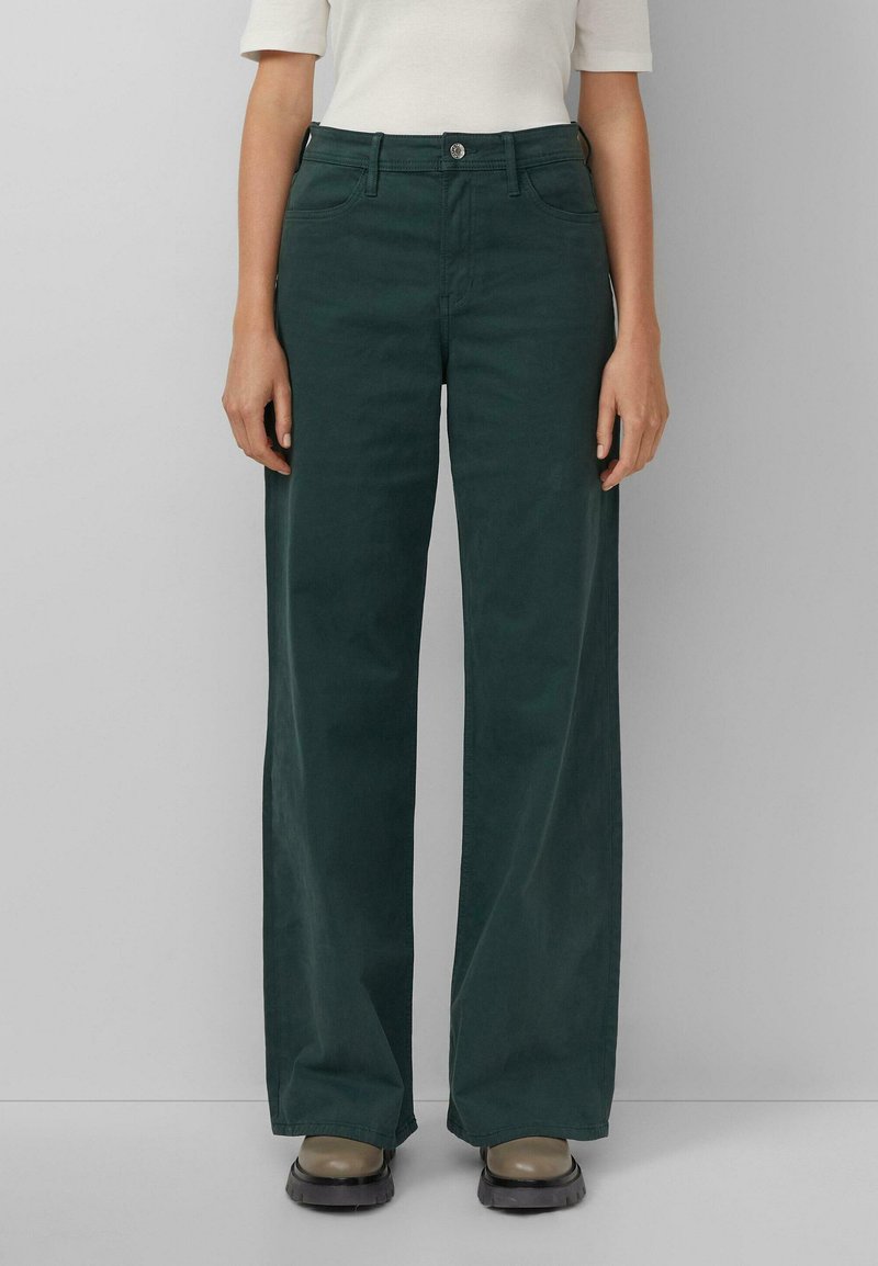 Dunkelgrüne, weit geschnittene Hose aus glattem Material mit einem Knopfverschluss vorne und Gürtelschlaufen. Kombiniert mit braunen Plateau-Schuhe.