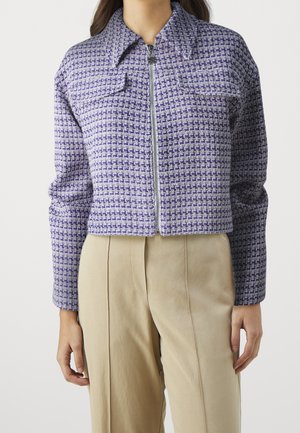 Nainen pukeutuneena lyhyeen violetti-valkoraidalliseen vetoketjulliseen takkiin, jossa on kaksi rintataskua, ja beigenvärisiin korkeavyötäröisiin housuihin.